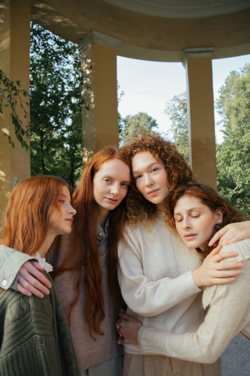 Four young women with red and curly hair embracing outdoors in a park.