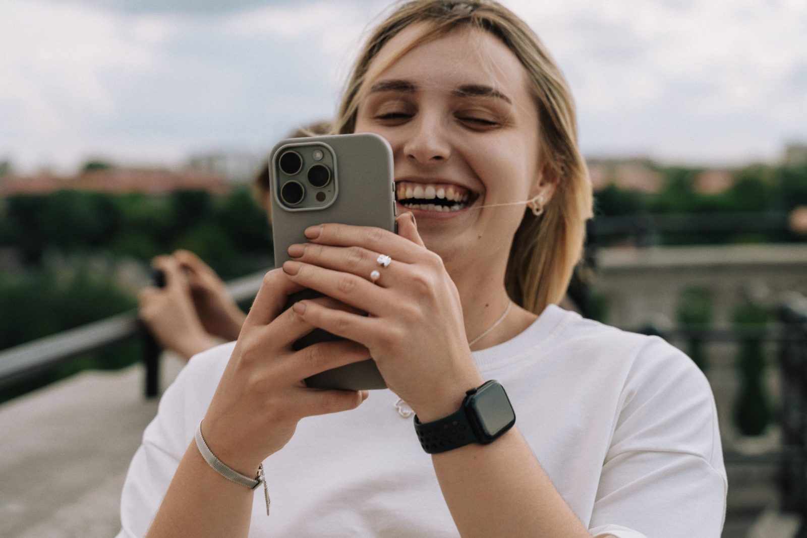A joyful young woman laughs while taking a selfie outside on a sunny day.