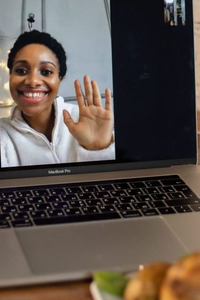 A smiling woman waves during a video call on a laptop, placed on a counter.